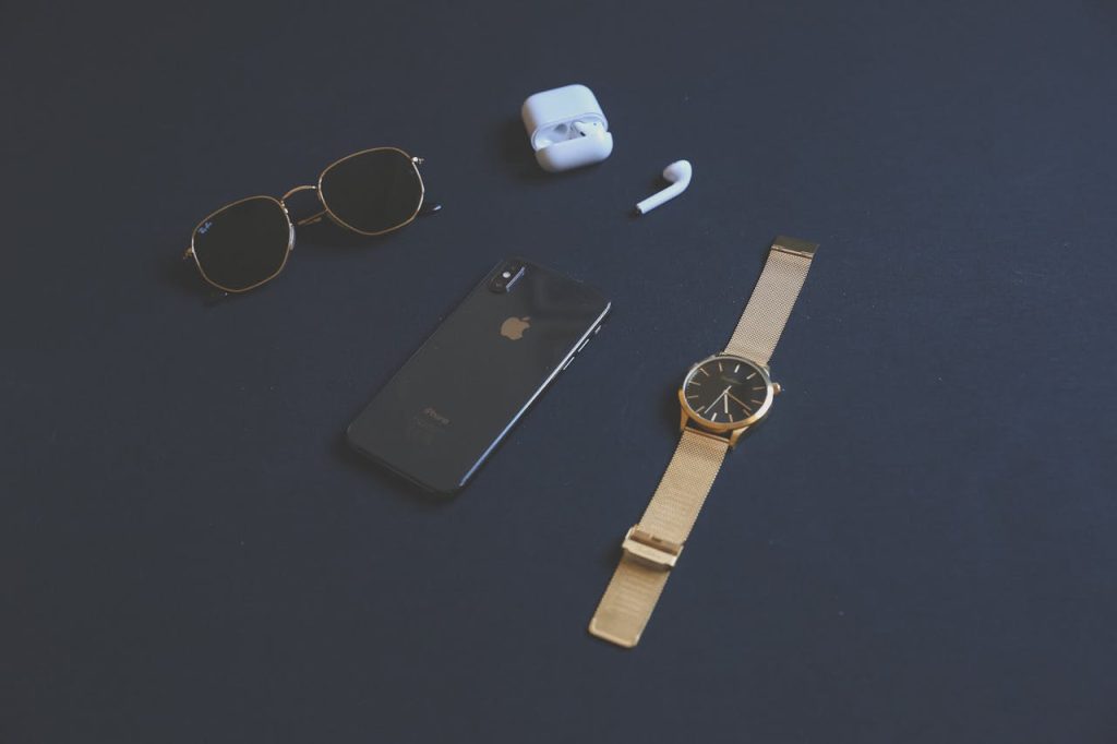 Flat lay of stylish accessories including a smartphone, gold watch, sunglasses, and earphones on a dark background.